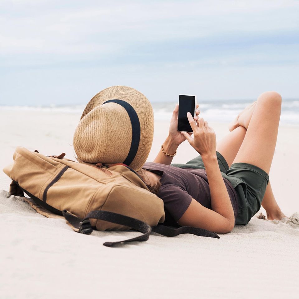 Katharina Weber: eine Frau liegt auf dem Rücken am Strand, Rucksack als Kopfkissen und Handy in der Hand
