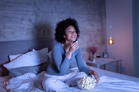 Ein junge Frau sitzt auf dem Bett und schaut mit Popcorn eine Serie