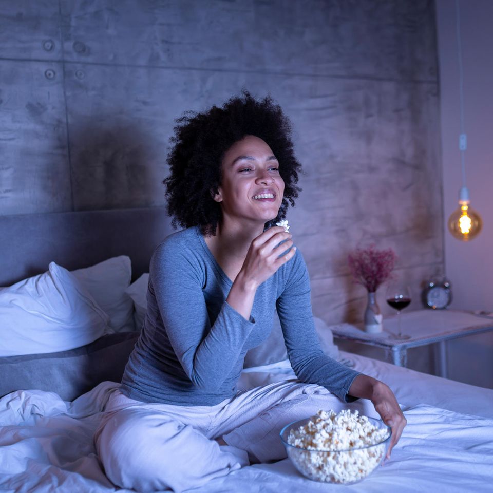 Ein junge Frau sitzt auf dem Bett und schaut mit Popcorn eine Serie