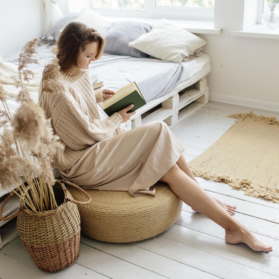 bruenette Frau in hellem Strickkleid sitzt auf einem Sitzkissen und liest in einem Buch