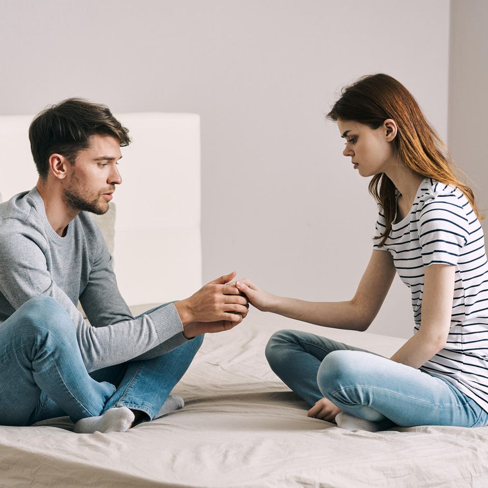 Emotionale Abhängigkeit lösen: Frau und Mann sitzen mit trauriger Miene auf einem Bett und halten sich an den Händen