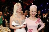 Dass zwischen Lady Gaga und Helen Mirren ein Altersunterschied von fast 40 Jahren liegt, würde man bei dem Strahlen der 76-Jährigen kaum vermuten. Die Schauspielerin setzt auf ein bodenlanges Kleid mit U-Ausschnitt und Blumen-Applikationen in Rosa, ihr Haar ziert ein ebenfalls rosafarbener Samt-Haarreifen. Lady Gaga trägt eine aufregende Robe in Weiß, ein integrierter Glitzer-BH sorgt für ein Wow-Dekolleté.