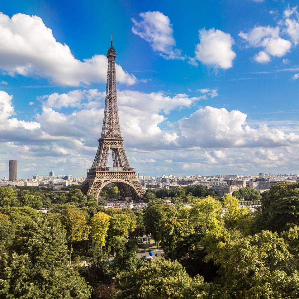 Paris: Grüne Bäume wachsen vor dem Eifelturm. Im Hintergrund ist der Himmel blau mit einigen weißen Wolken