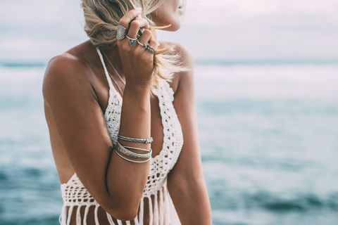 Eine blonde Frau steht am Strand und trägt Silberschmuck und ein Häkeltop