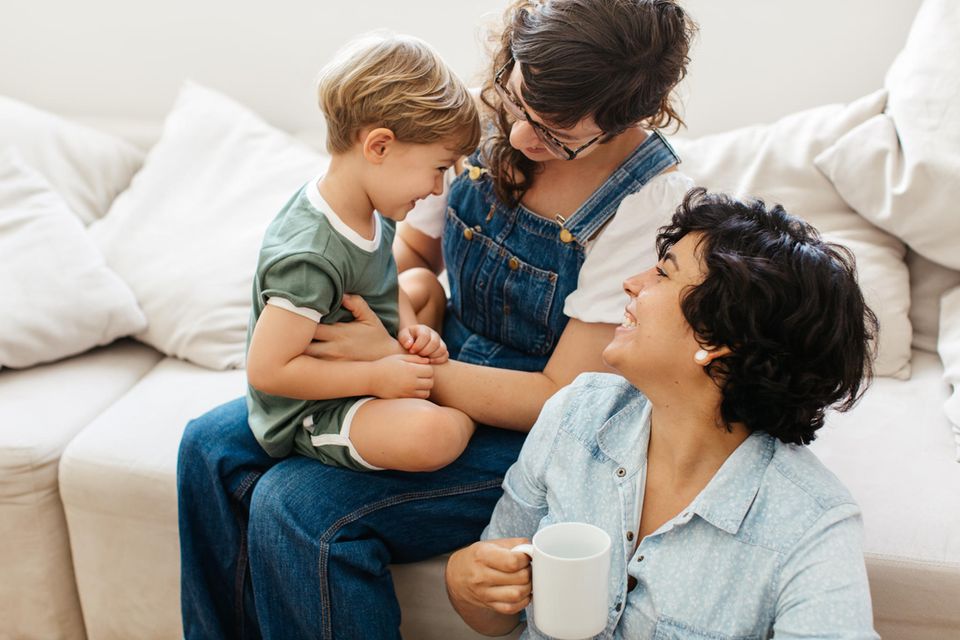 Single-Eltern: Ein Frau in Latzhose sitzt auf einem Sofa mit ihrem Sohn auf dem Schoß. Vor dem Sofa sitzt eine zweite Frau