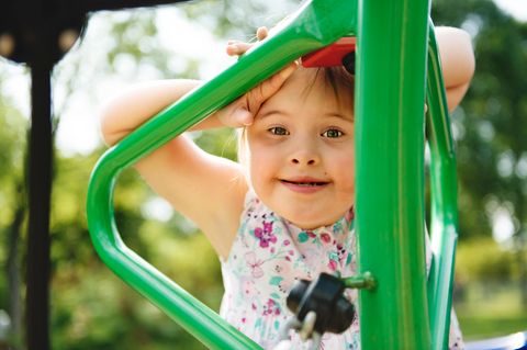 Barrierefreie Spielplätze: ein Mädchen mit Trisomie 21 lehnt an einem grünen Klettergerüst