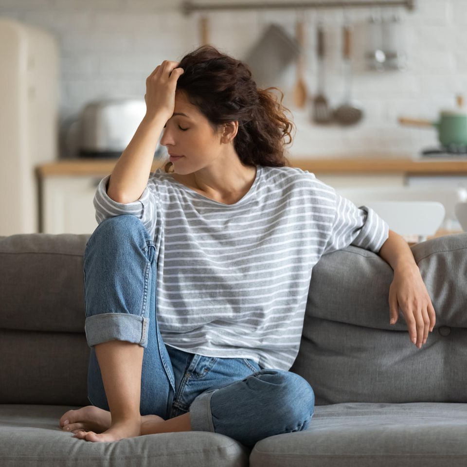 Frau sitzt auf Couch und hält sich Kopf.