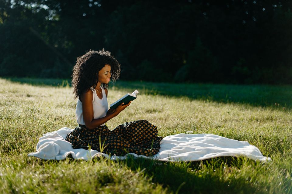 Frau mit Afro sitzt auf einer Wiese mit einem Enemies-to-Lovers Buch