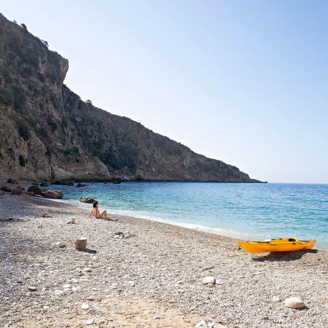 Albanien: eine Buch mit Felsen auf der linken Seite. Auf dem Sand sitzt eine Frau, neben ihr ein gelbes Kanu