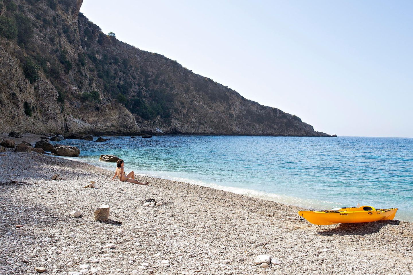 Albanien: eine Buch mit Felsen auf der linken Seite. Auf dem Sand sitzt eine Frau, neben ihr ein gelbes Kanu