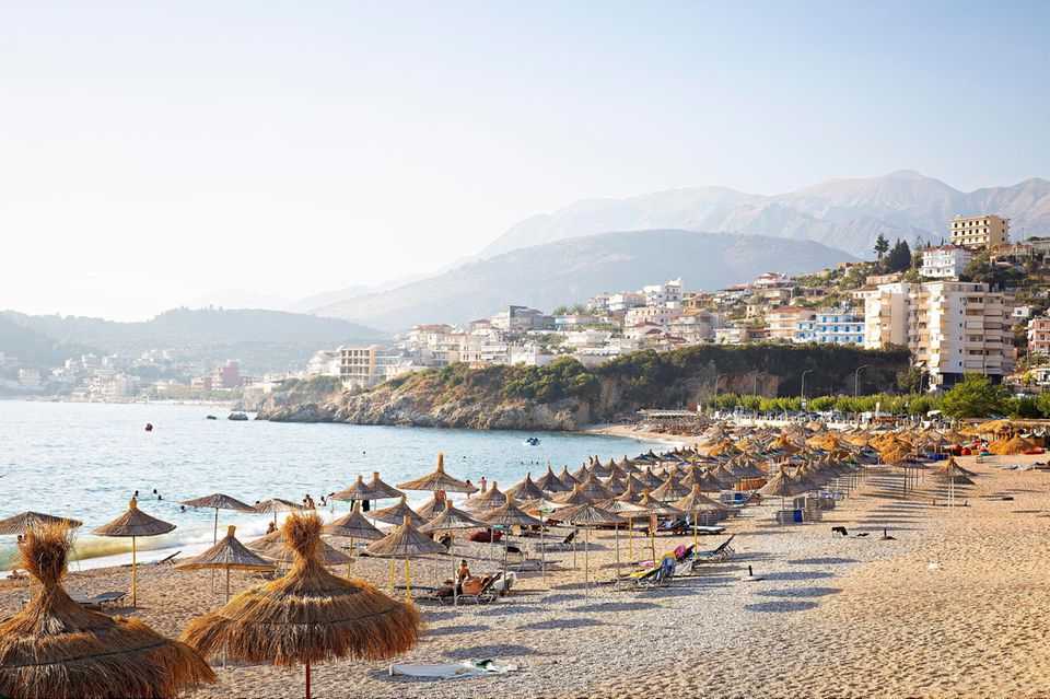 Albanien: ein Strand mit Strohsonnenschirmen. Im Hintergrund sind Hügel zu sehen, die im Dunst verschwinden