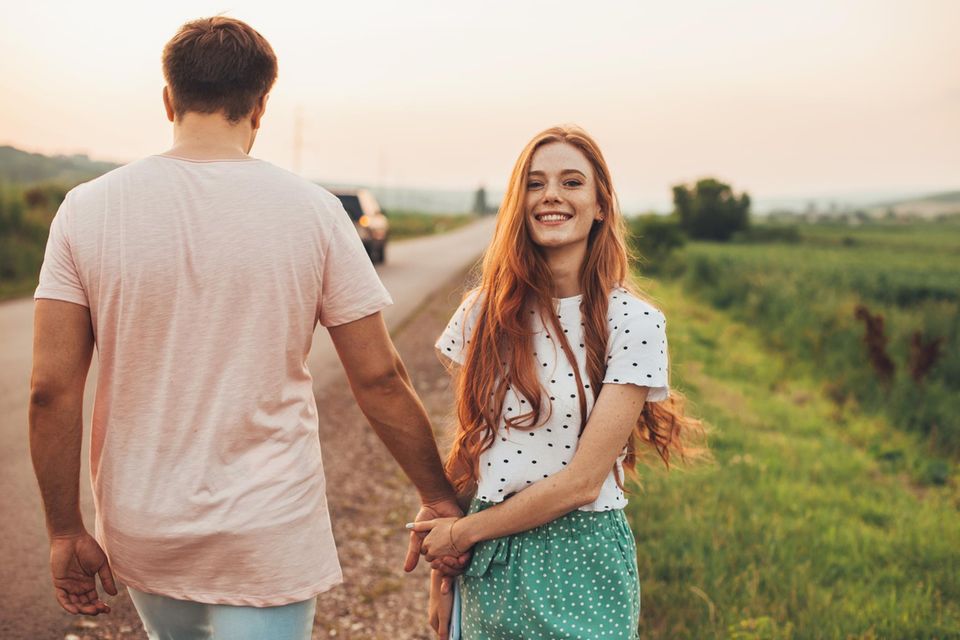 Lockere Beziehung: Frau hält lächelnd die Hand eines Mannes, der mit dem Rücken zugewandt steht