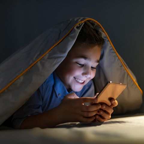 Junge liegt unter Decke und lacht in sein Smartphone.