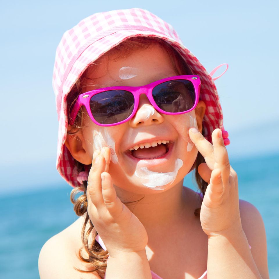 Mädchen hat Sonnenbrille auf der Nase, Sonnencreme im Gesicht und lacht fröhlich in die Kamera.