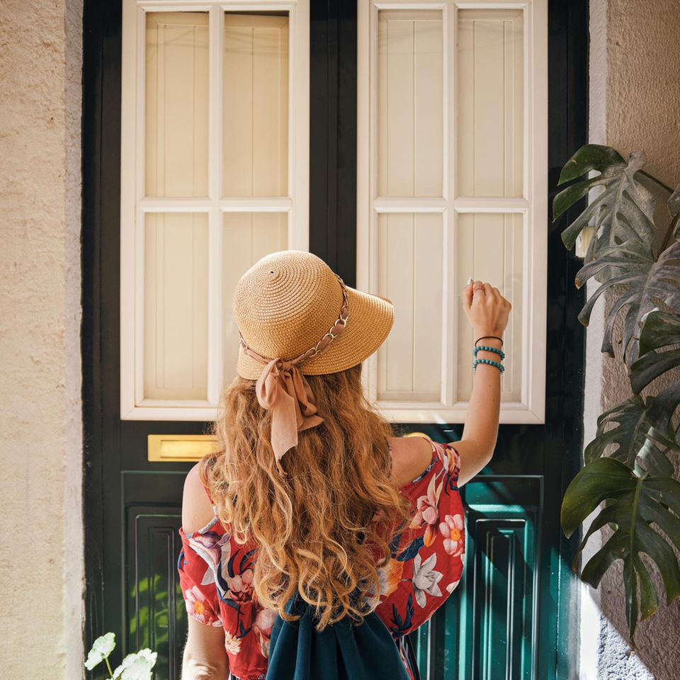 Andrea Lepperhoff: Eine Frau in sommerlicher Kleidung und Hut klopft an eine Haustür