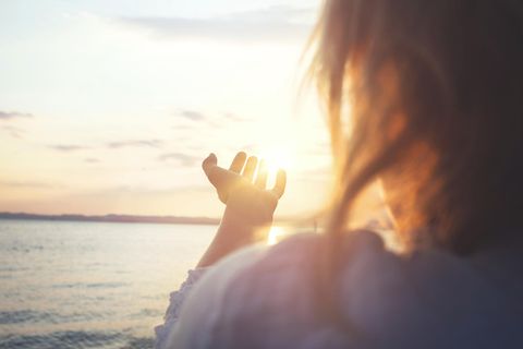Kleines Glück: Rückaufnahme einer Frau, die am Meer steht und die Hand nach der Sonne ausstreckt