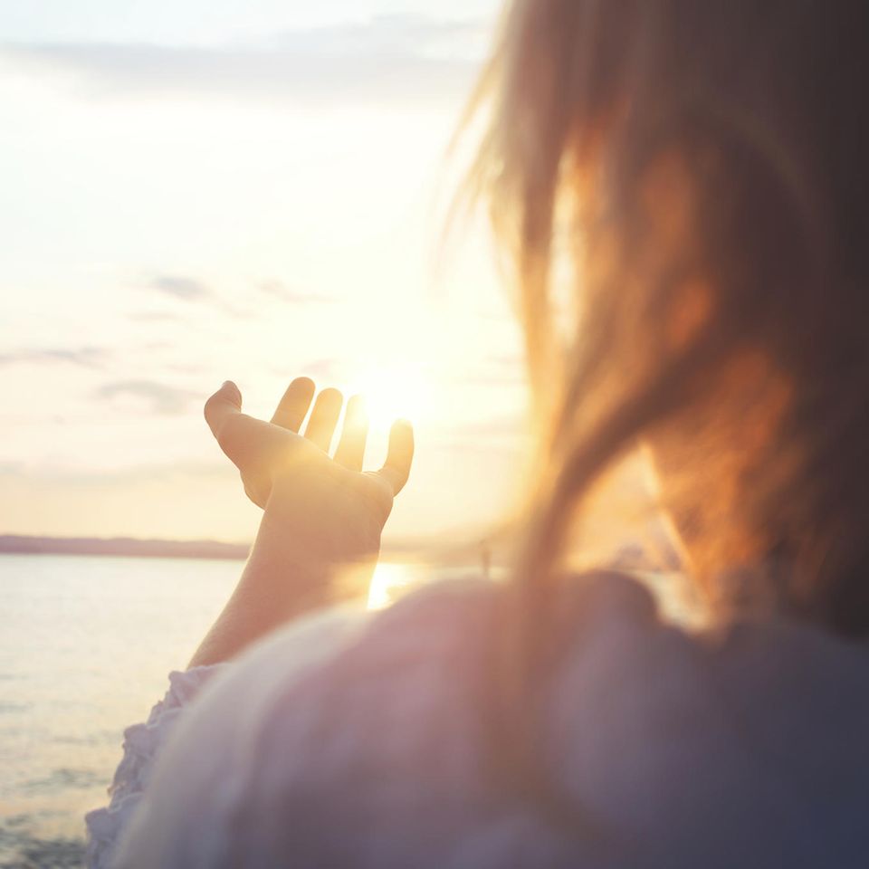 Kleines Glück: Rückaufnahme einer Frau, die am Meer steht und die Hand nach der Sonne ausstreckt