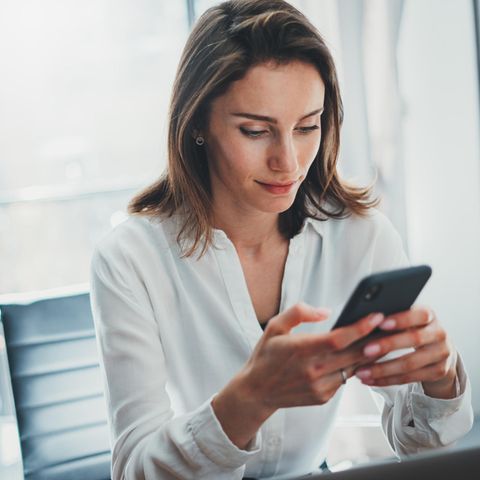 Finanz-Apps: eine Frau in weißer Bluse sitzt an einem Tisch mit einem Handy in der Hand