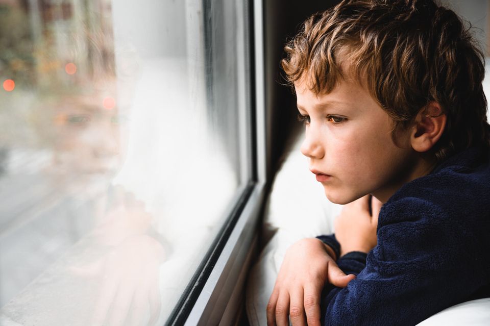 Junge sitzt am Fenster und schaut traurig nach draußen.
