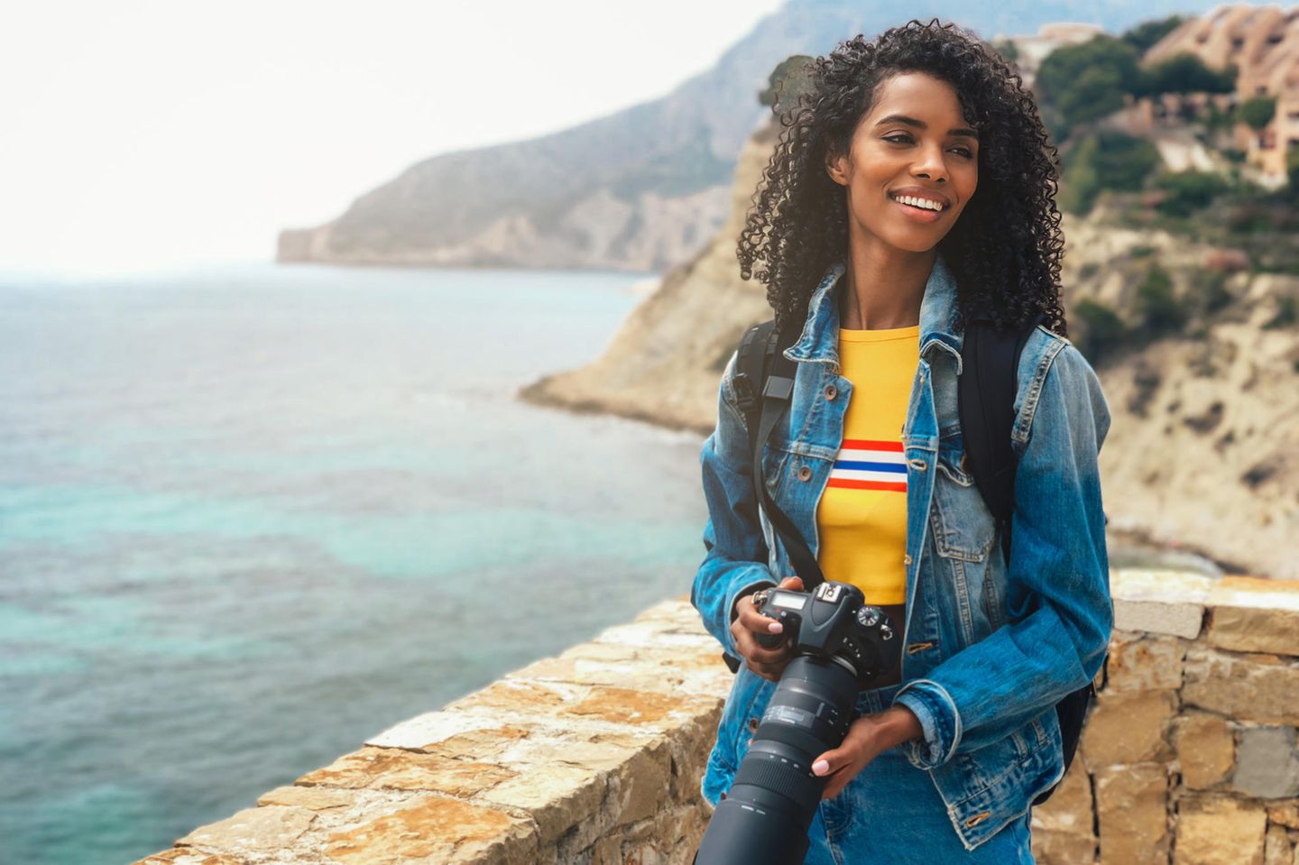 Frau mit Kamera am Meer: Deshalb macht Reisen nicht nur glücklich, sondern auch schlau
