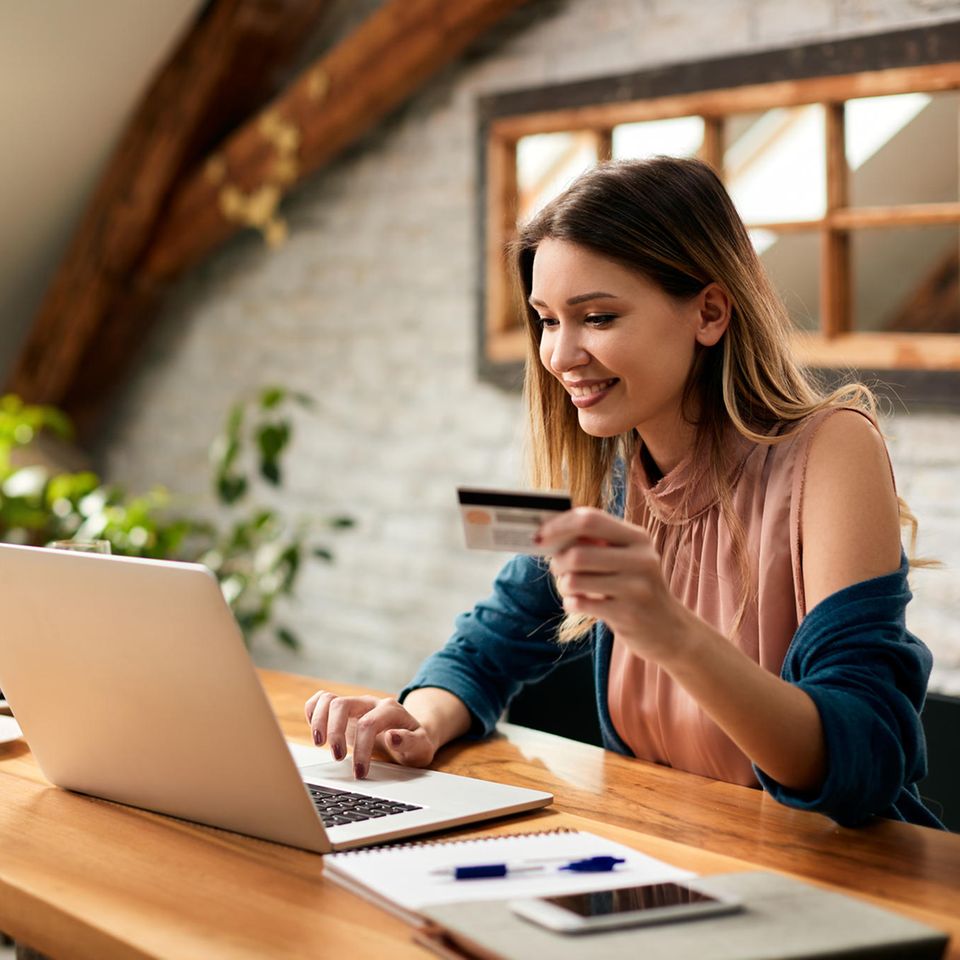 Eine blonde Frau sitzt an einem Laptop und hält in ihrer linken Hand eine Kreditkarte