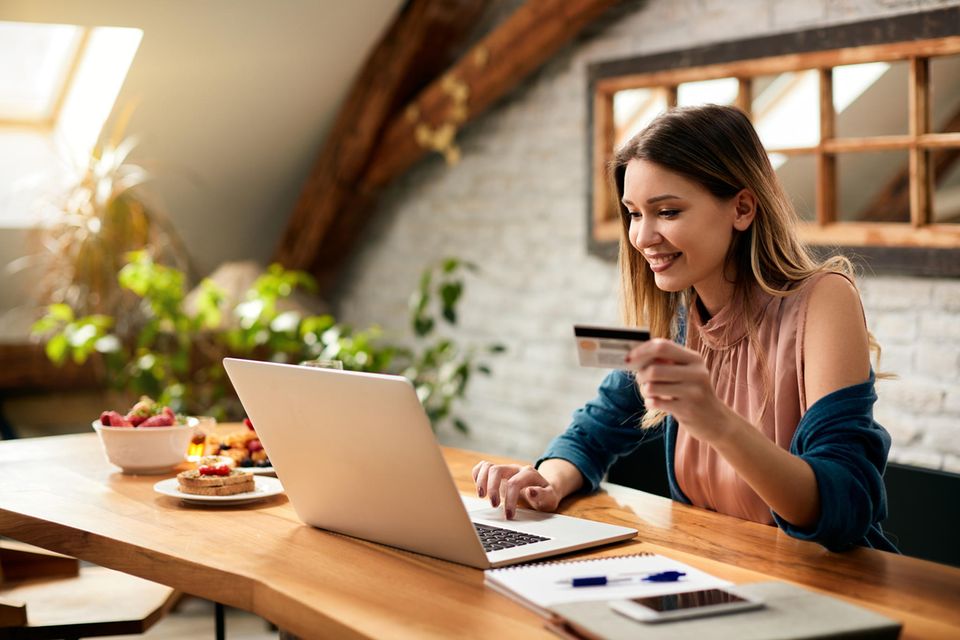 Eine blonde Frau sitzt an einem Laptop und hält in ihrer linken Hand eine Kreditkarte
