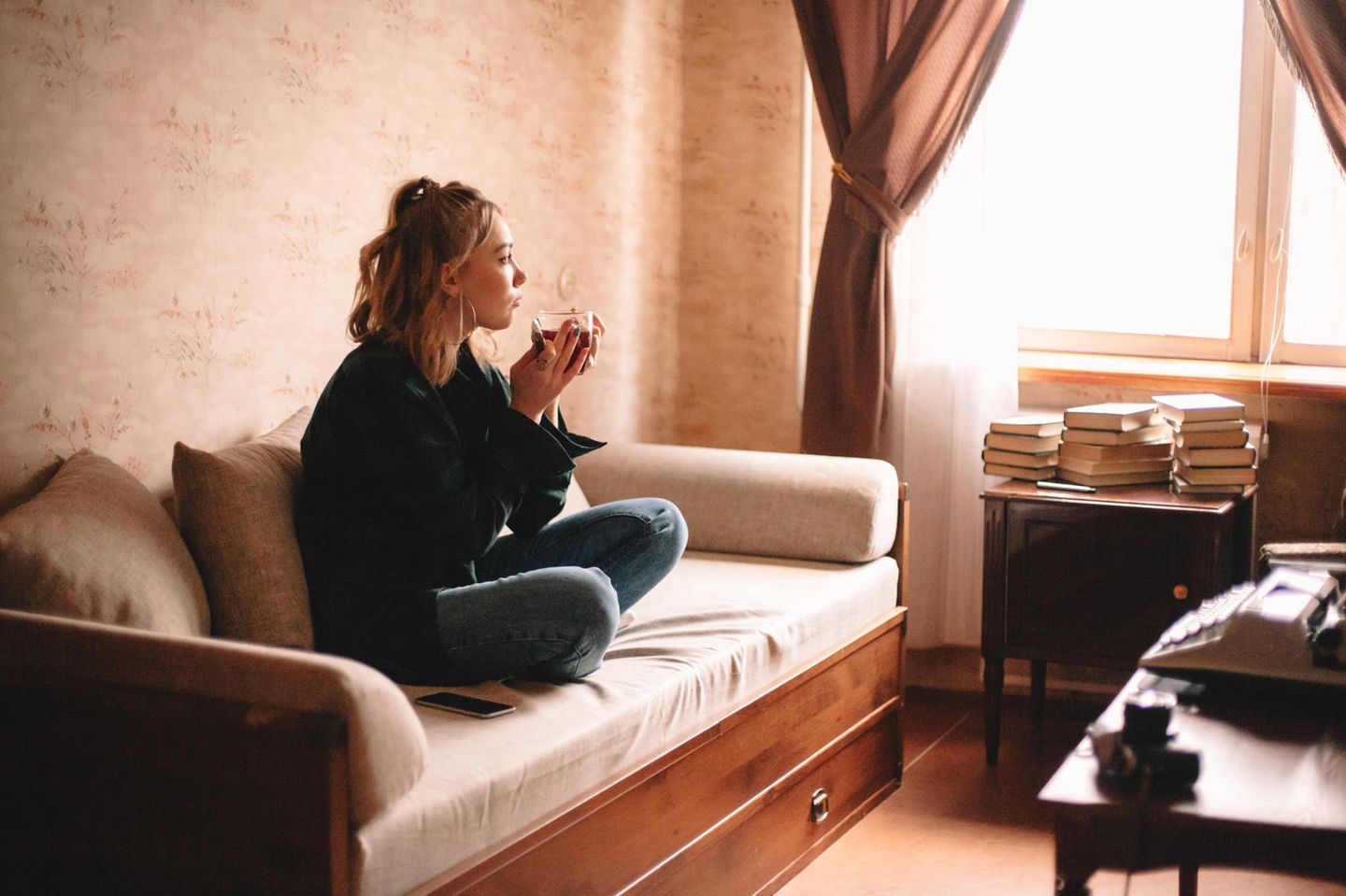 junge Frau hält eine Tasse Tee in den Händen, sitzt auf einem Sofa und blickt aus dem Fenster