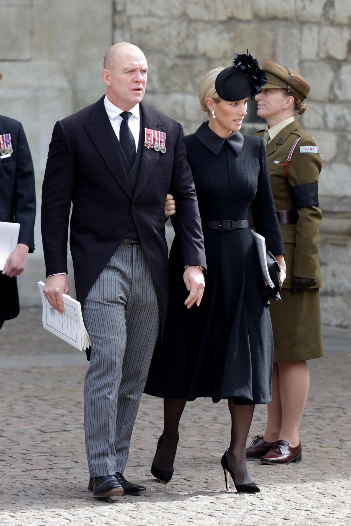 Zara Tindall trägt ein Mantelkleid von Laura Green, zu dem sie eine Maßschneiderung von Hutmacherin Juliette trägt. Wie andere Royals wählt auch Zara weißen Perlenschmuck. Diese Tradition ist auf Königin Victoria zurückzuführen, die nach dem Tod ihres Mannes nur noch Schwarz und Perlen getragen hat. 