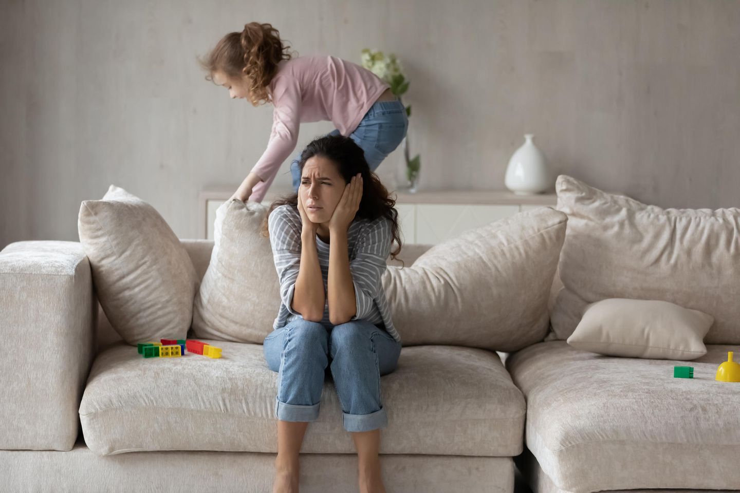 Familienkolumne: Mädchen turnt auf Sofa herum, während Mutter darauf sitzt und sich die Ohren zuhält