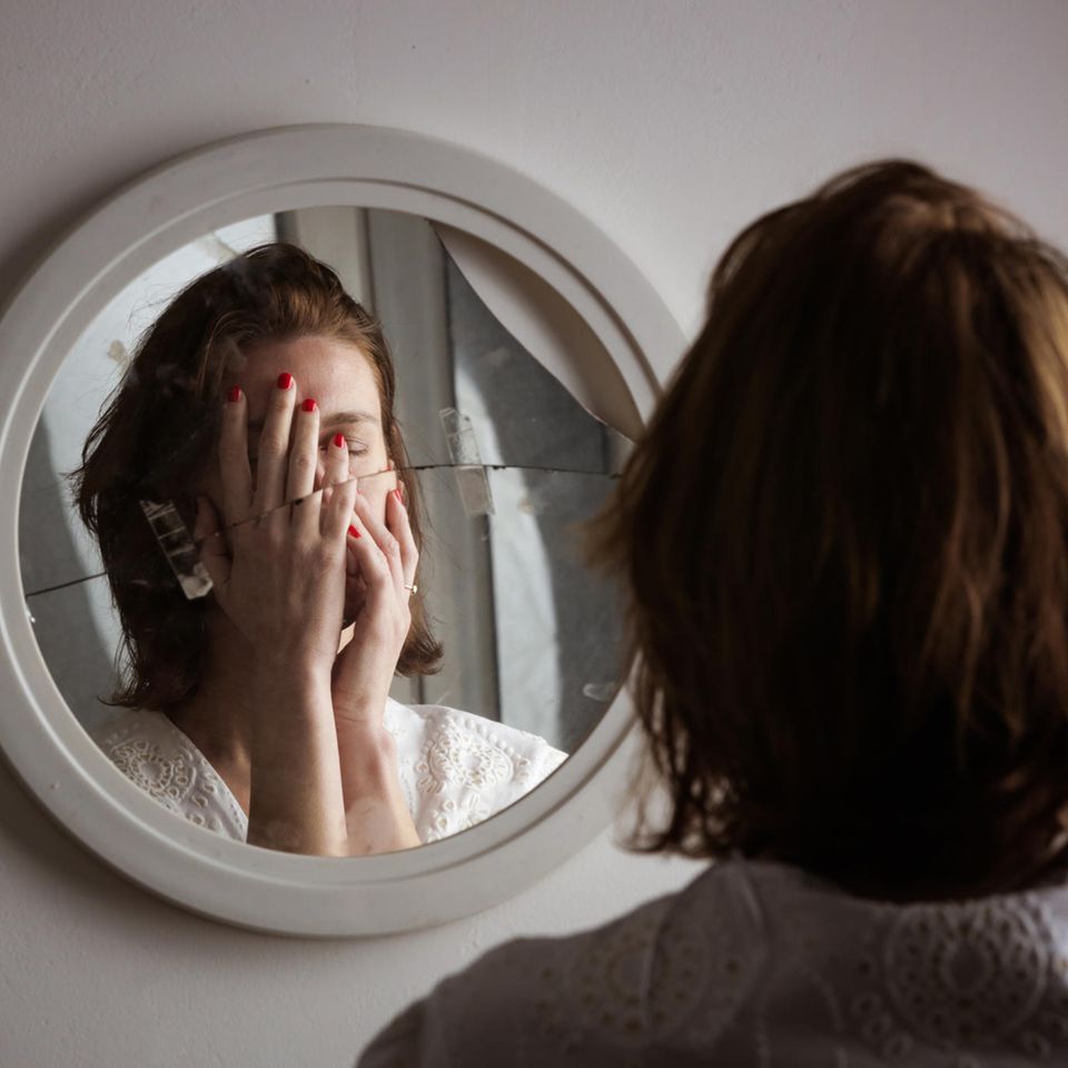 junge Frau steht vor einem zerbrochenen Spiegel und hält sich die Hände vor das Gesicht