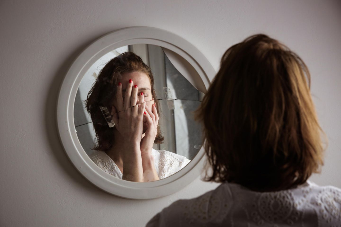 junge Frau steht vor einem zerbrochenen Spiegel und hält sich die Hände vor das Gesicht
