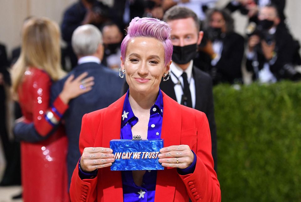 Megan Rapinoe trägt auf dem roten Teppich der Met Gala 2021 eine Clutch mit der Aufschrift "In gay we trust". Die Fußballspielerin setzt sich aktiv gegen Diskriminierung und Rassismus ein, was sie auch in ihrer Autobiografie verarbeitet, die sie 2021 geschrieben hat. 2012 bekannte sie sich öffentlich zu ihrer Homosexualität.