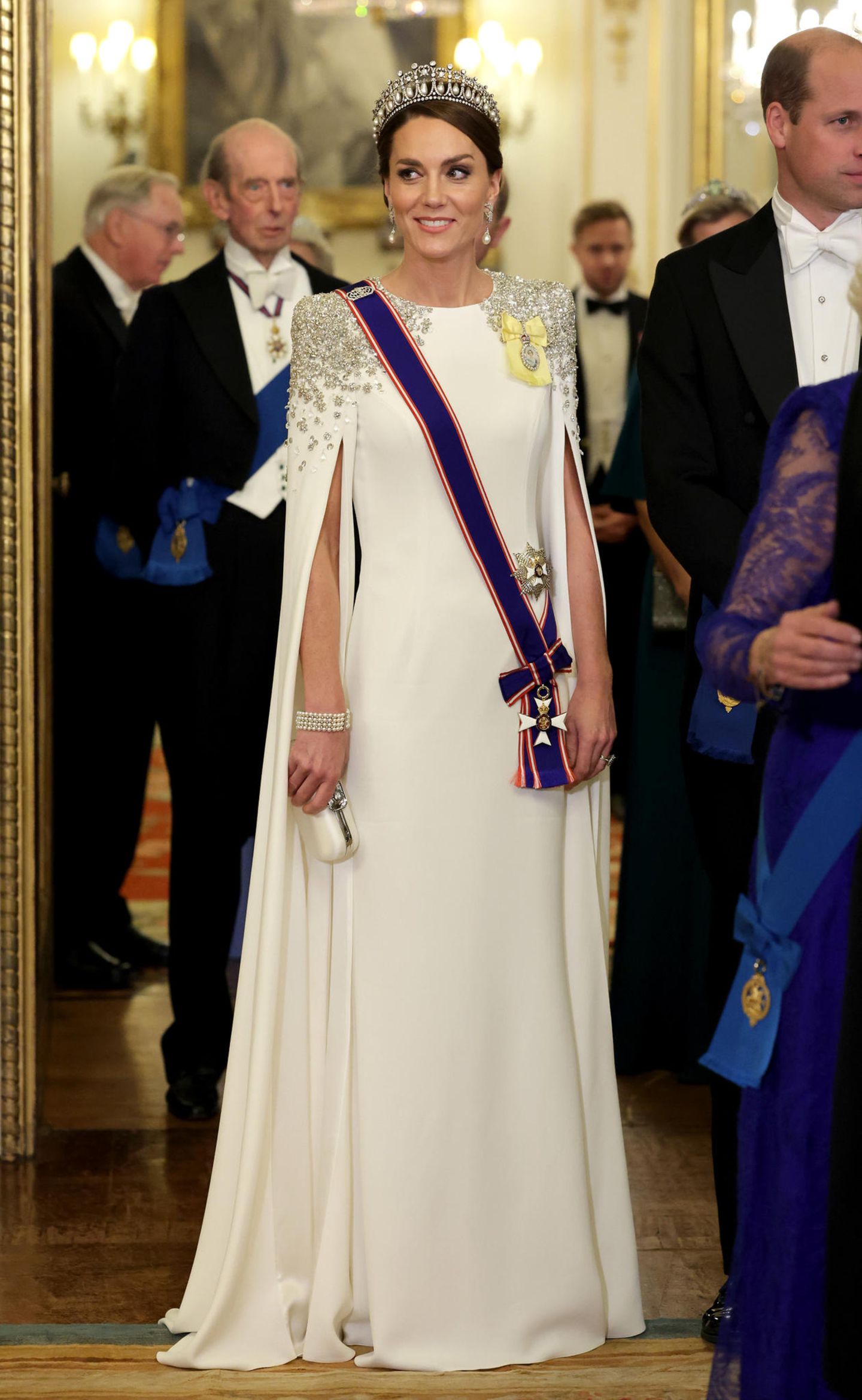 Beim ersten Staatsbankett während der Regentschaft von König Charles III. ist Catherine, Princess of Wales, der Hingucker des Abends. Die Wahl ihres Kleides und des Schmucks sind dabei besonders spannend. Denn, ihre bezaubernde Traumrobe in Weiß ist eigentlich ein Brautkleid. Es hat jedoch einen besonderen Kniff: An dem Kleid ihrer Lieblingsdesignerin Jenny Packham stechen besonders die aufwendige Schulterpartie mit Pailletten in Blumenform und die Cape-Ärmel hervor. Dazu wählt die Princess of Wales einen hochkarätigen Kopfschmuck: Die "Cambridge Lover's Knot" Tiara, welche ein Hochzeitsgeschenk für Prinzessin Diana war. Ihr luxuriöses Auftreten wird durch ein Armband von Queen Elizabeth II. ergänzt. 