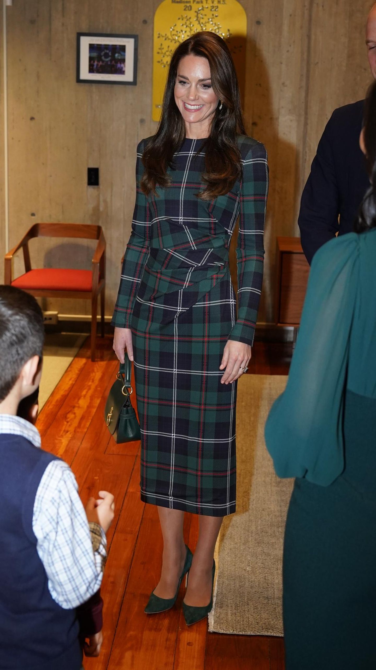 Catherine zieht sich im Laufe des Tages um und absolviert ihren zweiten Termin in einem neuen Tartan-Kleid von Burberry. Dazu stylt sie eine ihrer Lieblingstaschen, die Amberley Small Satchel Bag – ebenfalls in einer neuen grünen Variante – und passende smaragdfarbene Heels von Emmy London.