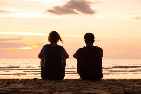 Zwei Personen sitzen am Strand vor einem Sonnenuntergang