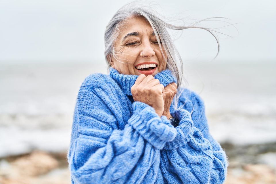 Eine ältere Frau steht glücklich in einem Winterpulli am Meer