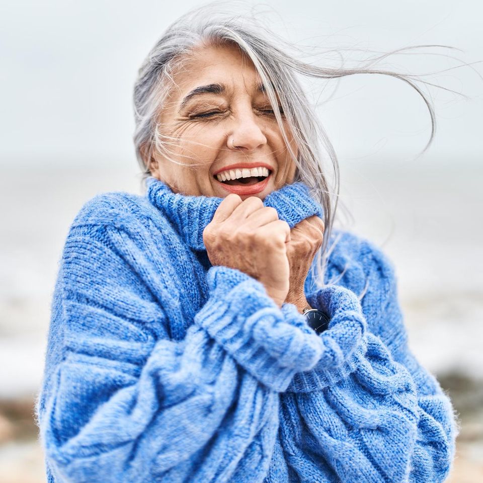 Eine ältere Frau steht glücklich in einem Winterpulli am Meer
