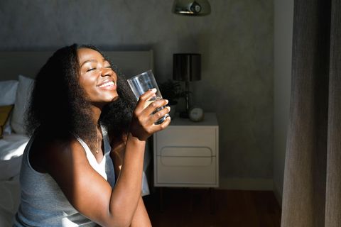 1%-Methode: Schöne Frau sitzt auf Bett mit einem Wasserglas und schaut in die Sonne