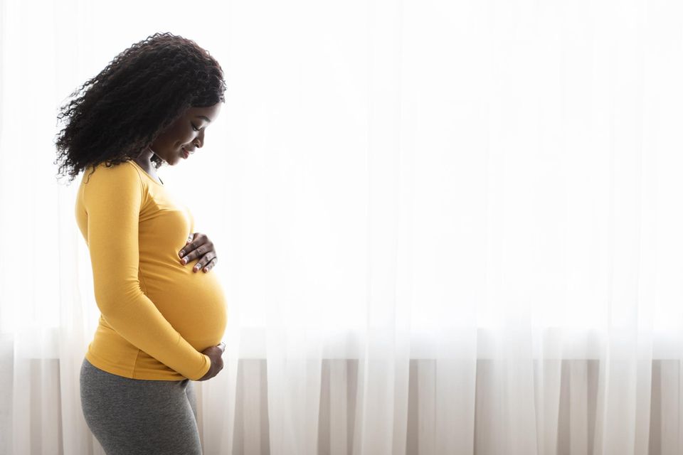 Hebammen klären auf: Eine schwangere junge Frau im gelben Oberteil umarmt lächelnd ihren Bauch
