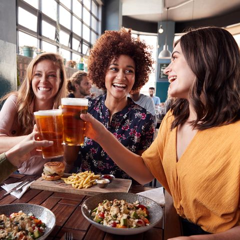 Vier junge Frauen essen gemeinsam Mittag und stoßen lachend mit Bier an