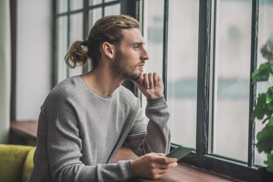 Die Phasen der Trennung bei Männern: Nachdenklicher Mann sitzt mit einem Handy in der Hand vor dem Fenster und schaut heraus
