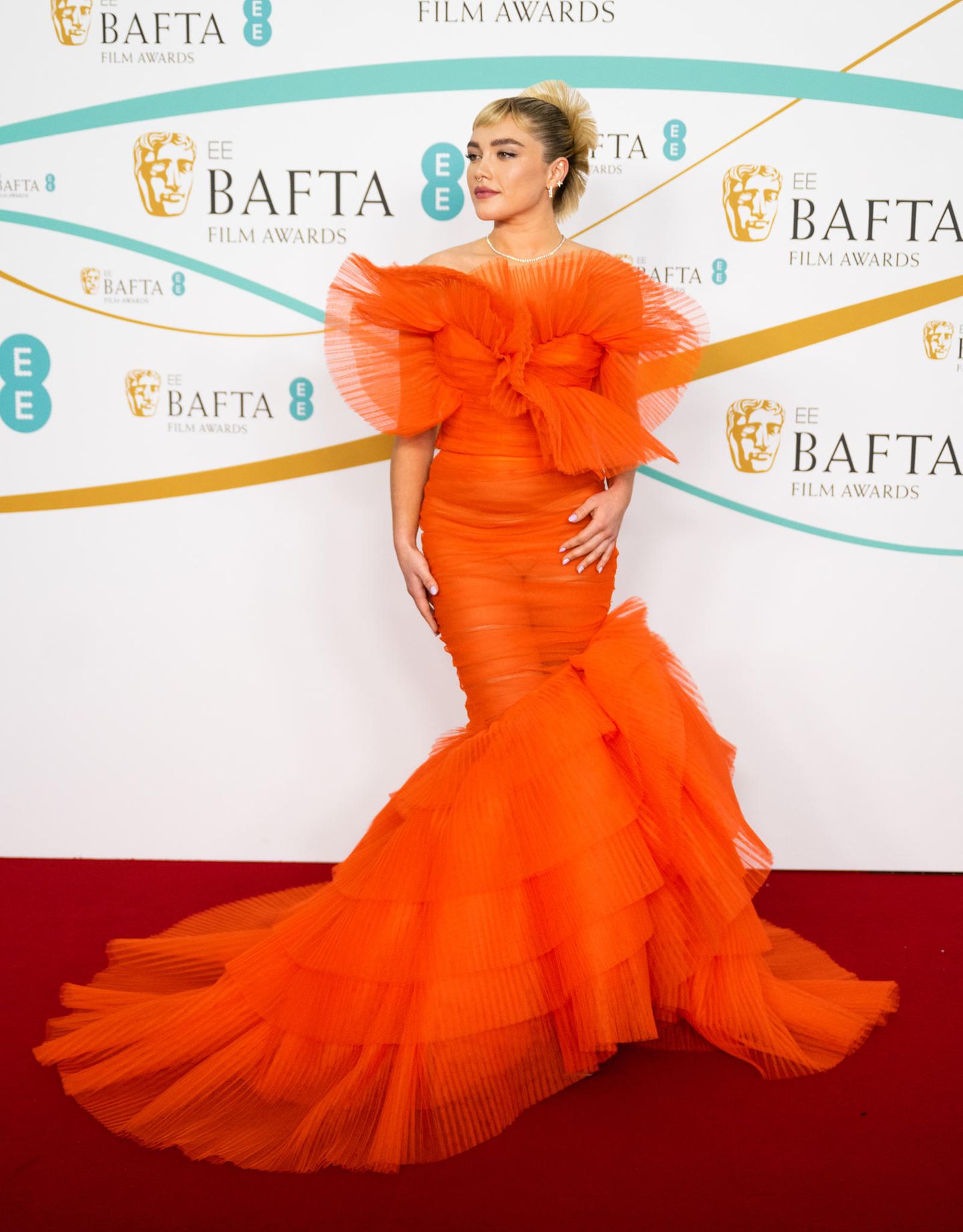 In London sorgt Florence Pugh in einem orangefarbenen transparenten Kleid von Nina Ricci für einen WOW-Moment. Ihr Haarstyle orientiert sich stark an ihrem Kleid und greift die Rüschen wieder auf. Diamanten-Schmuck und ein silberner Nasenring sorgen für einen interessanten Stilbruch.