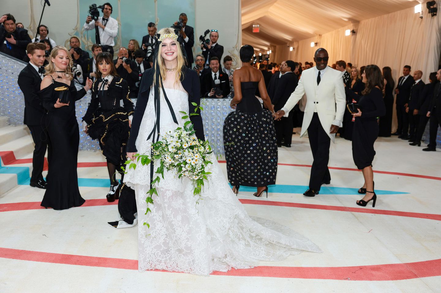 In ihrem weißen Spitzenkleid von Vivienne Westwood kombiniert mit Bolerojäckchen und Blumenschmuck aus Margeriten sieht Elle Fanning wirklich aus wie eine Maikönigin. Schmuck von Cartier macht diesen fröhlichen Look noch luxuriöser.