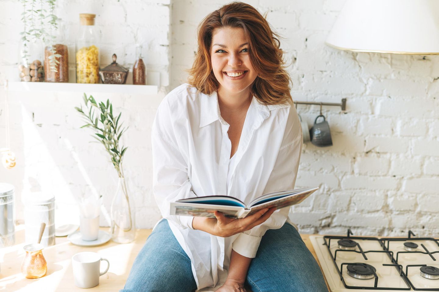 Brünette Frau lächelt, sitzt auf der Küchenarbeitsplatte und hält ein geöffnetes Kochbuch in den Händen