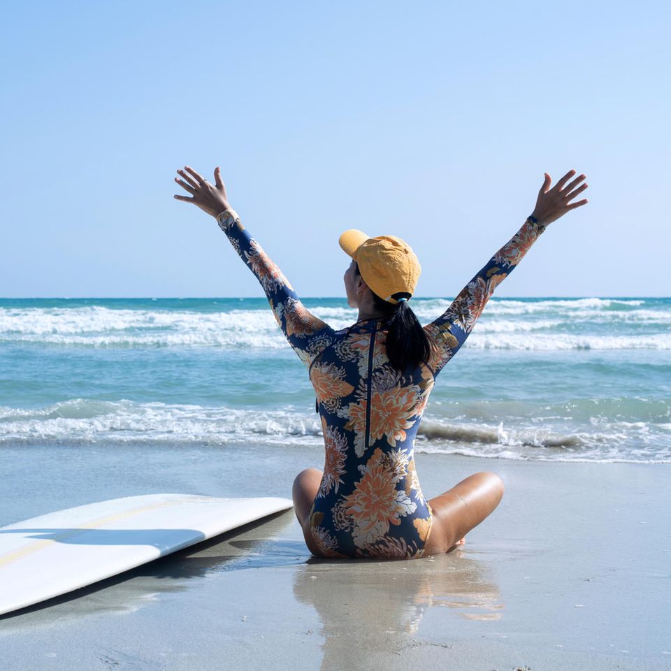 Surf-Badeanzug: Die perfekte Beachwear für einen aktiven Tag auf dem Wasser, Frau am Strand mit Surfbrett und Meer