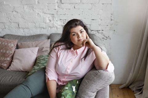 Abnehmspritze: Junge, hübsche, übergewichtige Frau sitzt nachdenklich auf dem Sofa