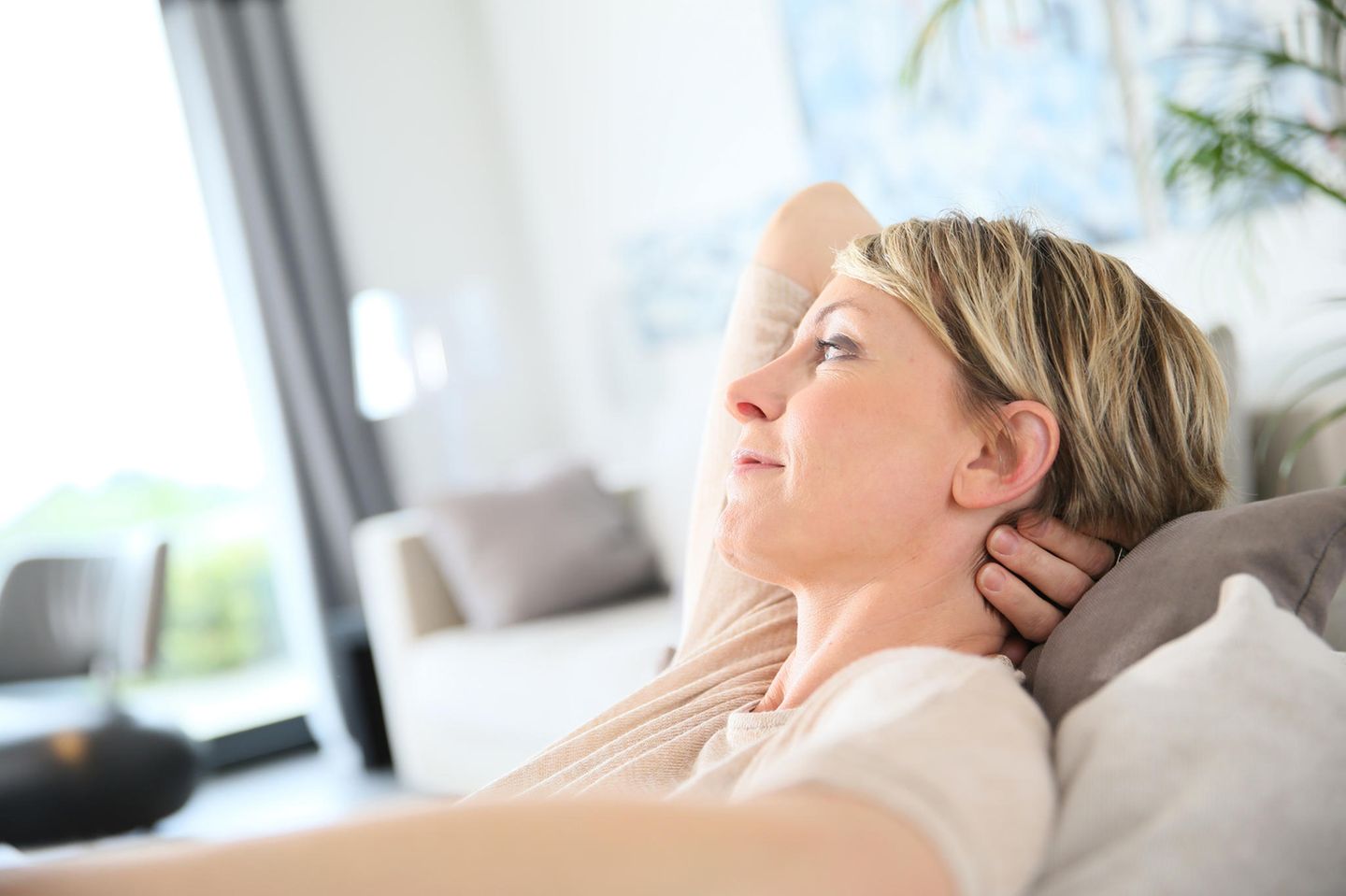 Prämenopause: Frau mit kurzen blonden Haaren sitzt zurückgelehnt auf dem Sofa, den Kopf auf dem Arm aufgestützt