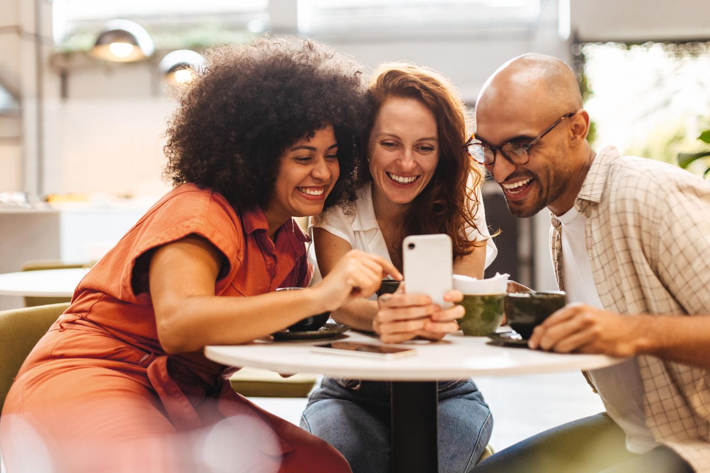 drei Freunde sitzen lachend an einem Tisch im Café und schauen gemeinsam auf ein Handy