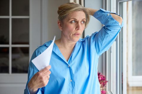 Perimenopause: Frau in blauer Bluse fächert sich mit Papier Luft zu und hält sich die Haare im Nacken hoch