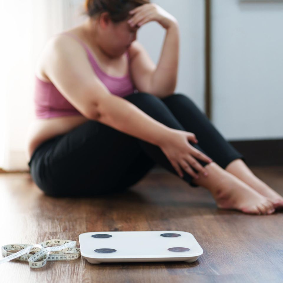 Kränkung durch Übergewicht: Frau sitzt traurig hinter Waage